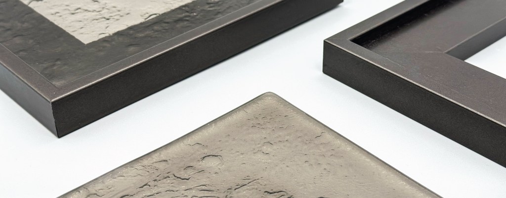 Close-up of bronze tinted artisan glass samples with textured kiln-formed finishes displayed alongside dark bronze aluminium framed door profiles manufactured by The Framed Door Co.